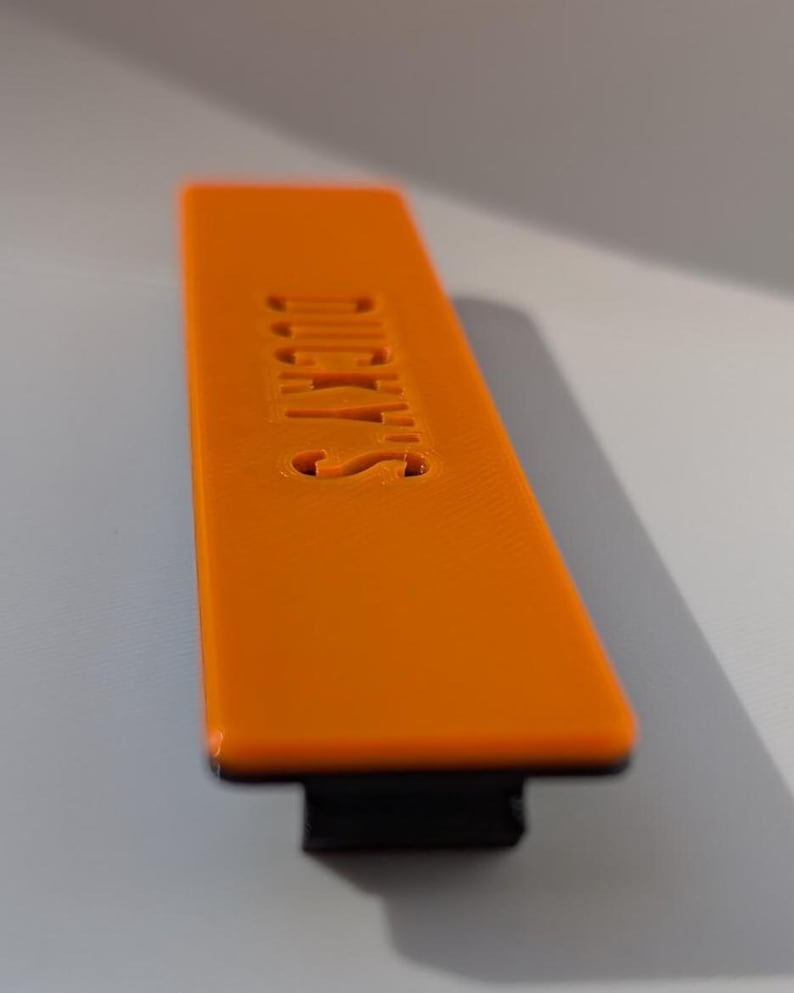 May include: An orange rectangular object with the word "DUCKY'S" embossed on its surface. The object has a black base and is set against a neutral background. The text is in a sans-serif font.