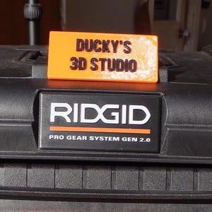 May include: An orange rectangular sign with the text "DUCKY'S 3D STUDIO" in black, resting on a black surface. Below, a black and white "RIDGID" logo with "PRO GEAR SYSTEM GEN 2.0" text. The image showcases a 3D printing studio.