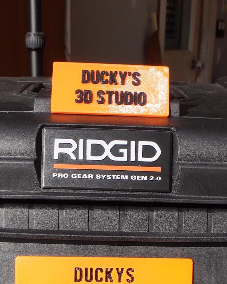May include: An orange rectangular sign with the text "DUCKY'S 3D STUDIO" in black, resting on a black surface. Below, a black and white "RIDGID" logo with "PRO GEAR SYSTEM GEN 2.0" text. The image showcases a 3D printing studio.