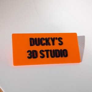 May include: An orange rectangular sign with the text "DUCKY'S 3D STUDIO" in black, raised lettering. The sign is set against a white background.