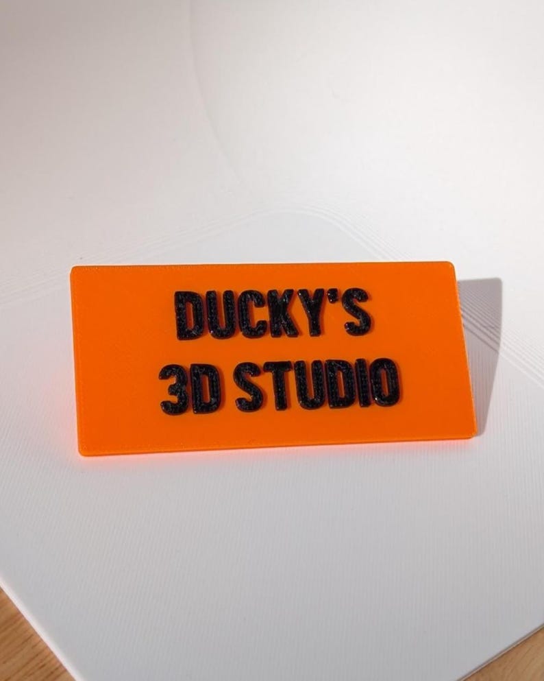 May include: An orange rectangular sign with the text "DUCKY'S 3D STUDIO" in black, raised lettering. The sign is set against a white background.