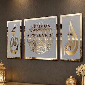 Peut inclure: Un ensemble de trois décorations murales rectangulaires présentant une calligraphie arabe en miroir doré sur un fond gris clair. Le panneau central carré affiche une écriture circulaire complexe, tandis que les deux panneaux latéraux présentent une calligraphie dorée verticale.