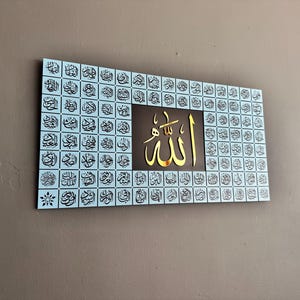 Puede incluir: Arte mural rectangular con caligraf&iacute;a &aacute;rabe. La obra de arte tiene un fondo azul claro con numerosos cuadrados de escritura negros. Un cuadrado central marr&oacute;n muestra la palabra "Allah" en color dorado.