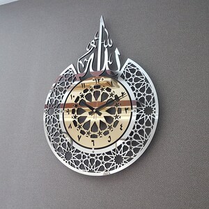 May include: A silver and gold wall clock with an intricate geometric design and the Arabic word "Allah" in the center. The clock face features black Roman numerals and a black hour and minute hand.