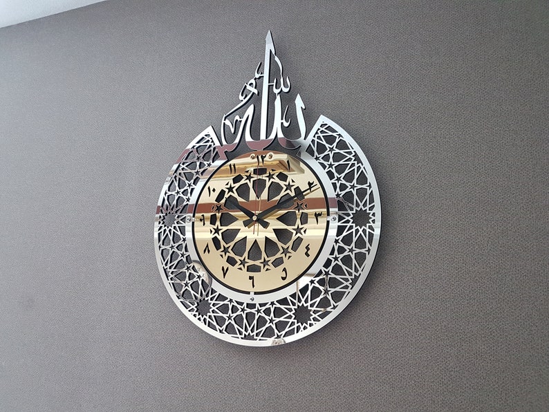 May include: A silver and gold wall clock with an intricate geometric design and the Arabic word "Allah" in the center. The clock face features black Roman numerals and a black hour and minute hand.