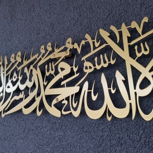 Może przedstawiać: Złote metalowe dzieło sztuki ściennej z arabską kaligrafią. Grafika jest zamontowana na ciemnoniebieskiej, teksturowanej powierzchni. Pismo jest elegancko zaprojektowane i wydaje się być tekstem religijnym lub duchowym.