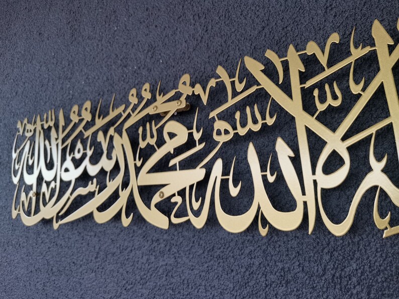 Może przedstawiać: Złote metalowe dzieło sztuki ściennej z arabską kaligrafią. Grafika jest zamontowana na ciemnoniebieskiej, teksturowanej powierzchni. Pismo jest elegancko zaprojektowane i wydaje się być tekstem religijnym lub duchowym.
