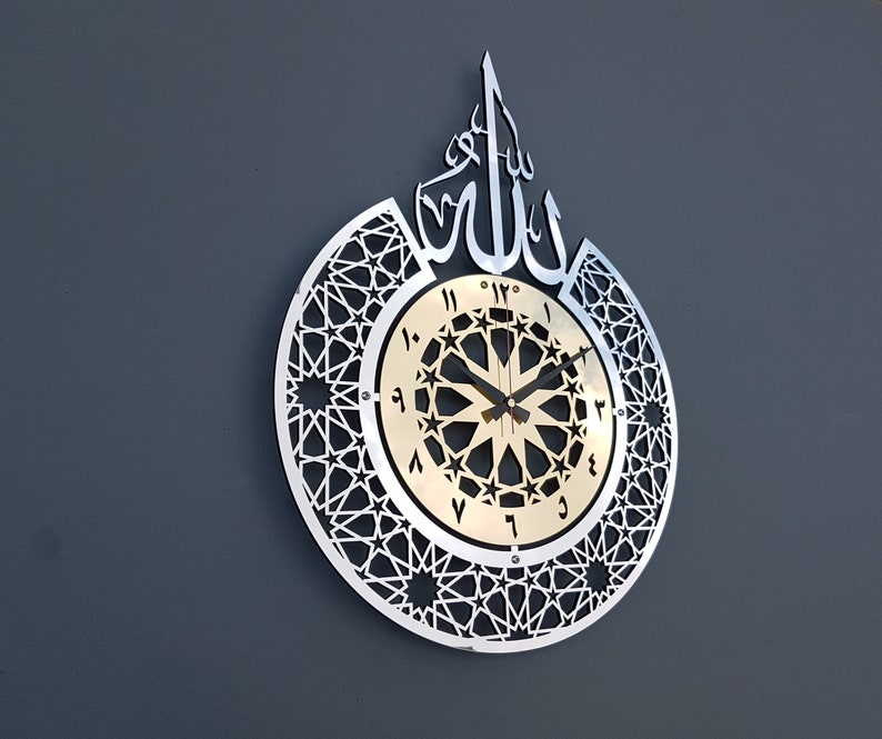 May include: A silver and gold wall clock with an intricate geometric design and Arabic calligraphy. The clock face features a gold background with black Roman numerals and a black star pattern.