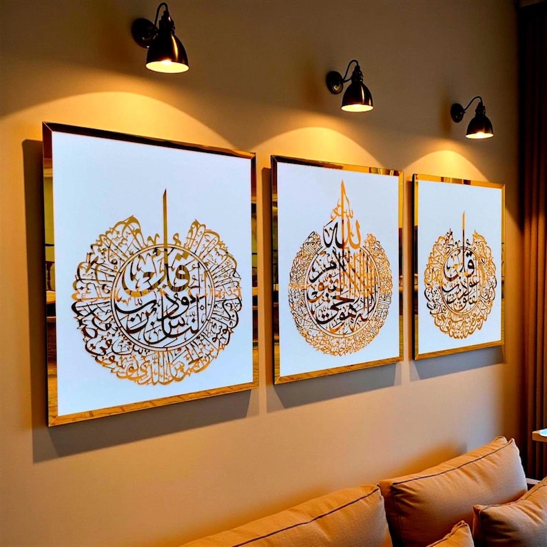 May include: Three framed wall art pieces with gold calligraphy on a white background. The frames have a gold border. The art is displayed on a tan wall, with three black sconce lights above. The art is above a tan couch.