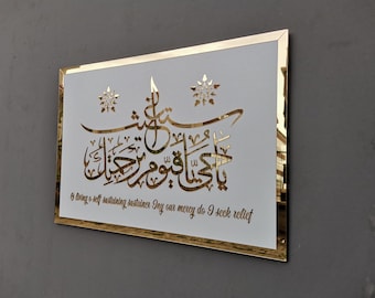 May include: A white rectangular wall art piece with a gold frame. The art features a gold Arabic calligraphy design with the text "يا حيا قيوم رحيل" and the English translation "& bring o self sustaining sustainer Iny our mercy do I seck relief".