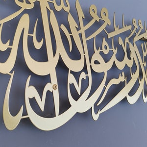 Może przedstawiać: Złote metalowe dzieło sztuki ściennej z arabską kaligrafią. Grafika jest zamontowana na szarej ścianie i przedstawia skomplikowany skrypt o dekoracyjnej estetyce. Metal ma matowe wykończenie.