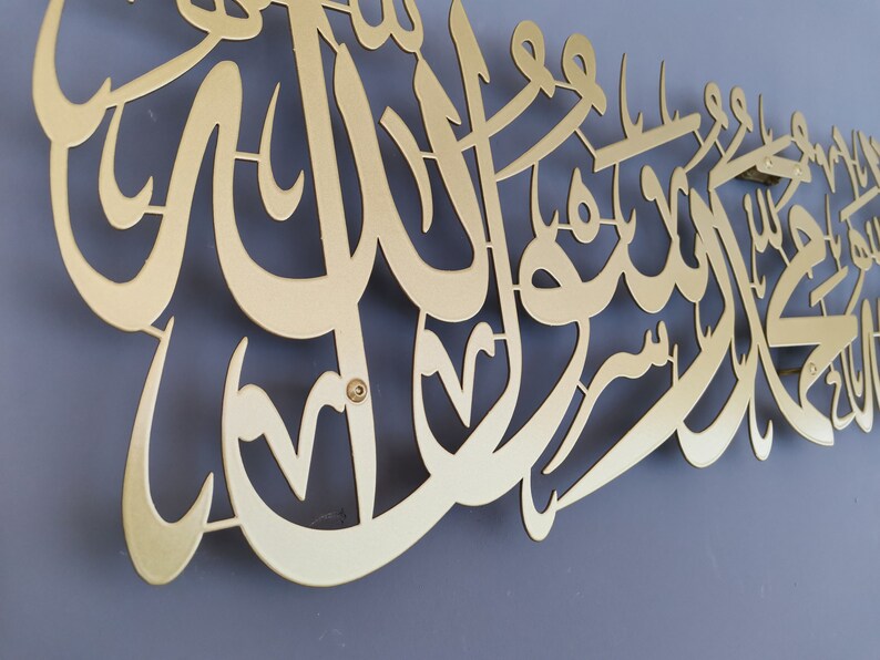 Może przedstawiać: Złote metalowe dzieło sztuki ściennej z arabską kaligrafią. Grafika jest zamontowana na szarej ścianie i przedstawia skomplikowany skrypt o dekoracyjnej estetyce. Metal ma matowe wykończenie.