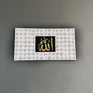 Puede incluir: Arte mural rectangular con caligraf&iacute;a dorada de la palabra "Allah" sobre un fondo negro. La obra de arte est&aacute; enmarcada por una cuadr&iacute;cula de peque&ntilde;os cuadrados de escritura &aacute;rabe en blanco y negro. Se exhibe contra una pared gris.