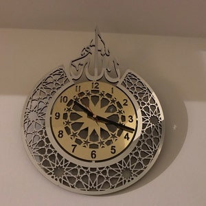 May include: A silver and gold wall clock with an intricate geometric design. The clock face features Arabic numerals and a starburst pattern. The clock is shaped like a crescent moon with a stylized Arabic calligraphy design at the top.