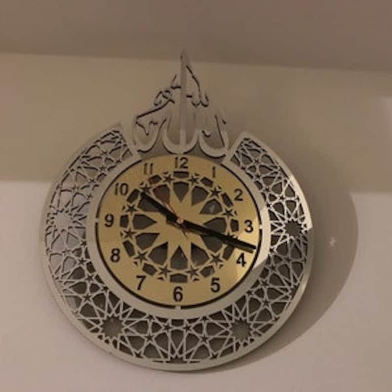 May include: A silver and gold wall clock with an intricate geometric design. The clock face features Arabic numerals and a starburst pattern. The clock is shaped like a crescent moon with a stylized Arabic calligraphy design at the top.