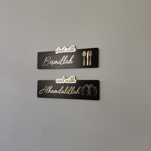 May include: Two black rectangular signs with gold lettering and icons. The top sign reads "start with Bismillah" with a gold fork, knife, and spoon. The bottom sign reads "end with Alhamdulillah" with two gold hands in a praying position.