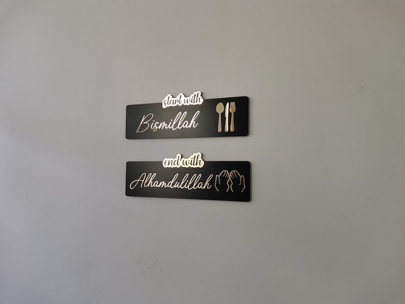 May include: Two black rectangular signs with gold lettering and icons. The top sign reads "start with Bismillah" with a gold fork, knife, and spoon. The bottom sign reads "end with Alhamdulillah" with two gold hands in a praying position.