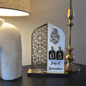 May include: A white wooden Ramadan countdown calendar with a gold geometric design. The calendar features a black number "03" and the text "Day of Ramadan".