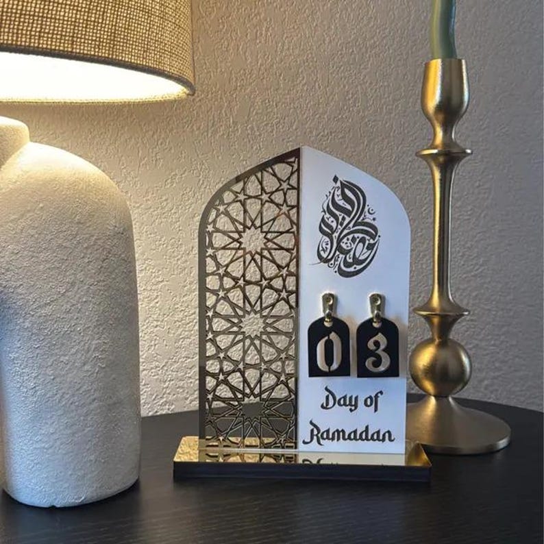 May include: A white wooden Ramadan countdown calendar with a gold geometric design. The calendar features a black number "03" and the text "Day of Ramadan".