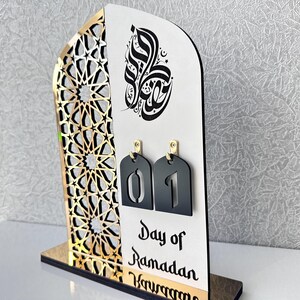 May include: A white and gold Ramadan countdown calendar with black numbers. The calendar features an intricate geometric design and Arabic calligraphy. The text reads "Day of Ramadan".