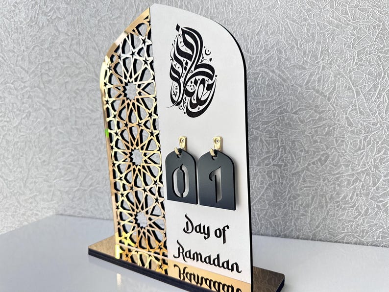May include: A white and gold Ramadan countdown calendar with black numbers. The calendar features an intricate geometric design and Arabic calligraphy. The text reads "Day of Ramadan".