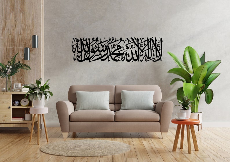Może przedstawiać: Czarna metalowa grafika ścienna z arabską kaligrafią, zamontowana nad beżową sofą. Grafika to długi, poziomy wz&oacute;r ze skomplikowanym pismem. Pok&oacute;j jest ozdobiony roślinami, drewnianą szafką i małym okrągłym dywanem.