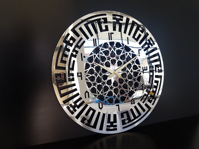 May include: A silver and black wall clock with Arabic calligraphy around the edge and a black and white geometric design in the center. The clock hands are gold.