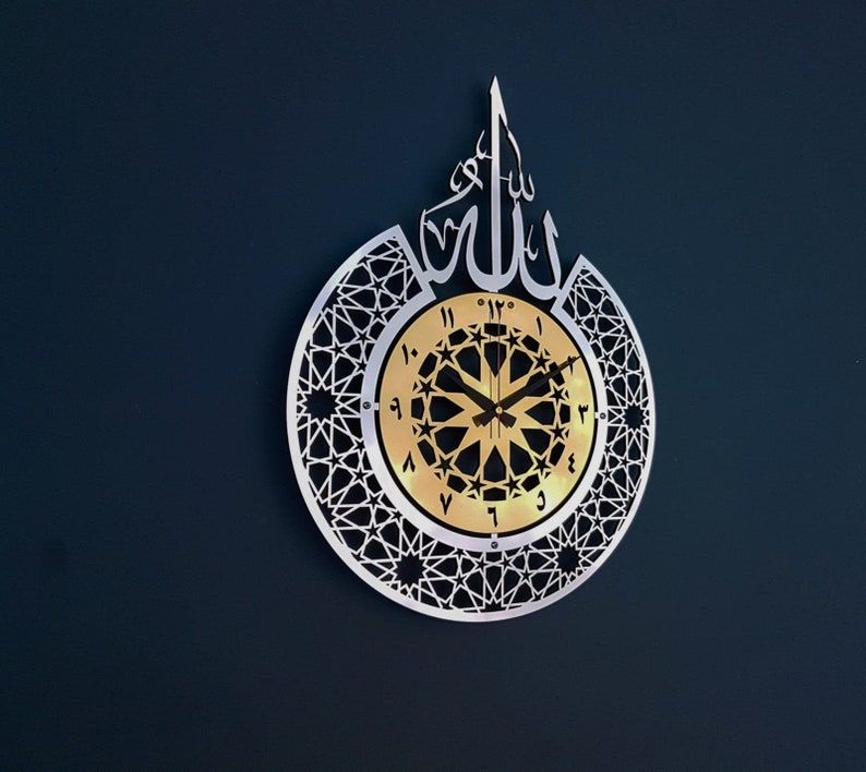 May include: A silver wall clock with an intricate geometric design and Arabic numerals. The clock face features a gold background with a black starburst pattern and black hands. The word "Allah" is written in Arabic script at the top of the clock.