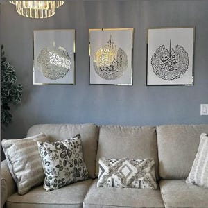 May include: Three framed art pieces with gold calligraphy on a white background. The frames have a gold border and are mounted on a gray wall above a beige sofa with decorative pillows. A chandelier hangs above the art.