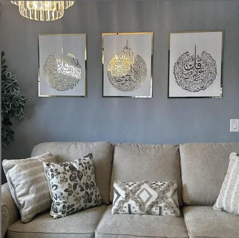 May include: Three framed art pieces with gold calligraphy on a white background. The frames have a gold border and are mounted on a gray wall above a beige sofa with decorative pillows. A chandelier hangs above the art.