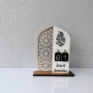 May include: A white wooden Ramadan calendar with a gold geometric pattern and Arabic calligraphy. The calendar has a black number tag with the number "01" on it. The text "Day of Ramadan" is printed below the number tag.