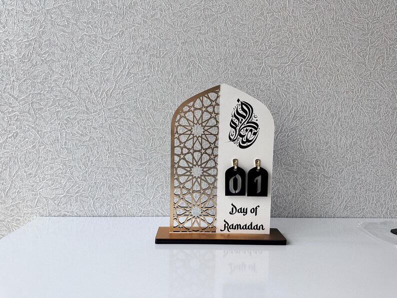 May include: A white wooden Ramadan calendar with a gold geometric pattern and Arabic calligraphy. The calendar has a black number tag with the number "01" on it. The text "Day of Ramadan" is printed below the number tag.