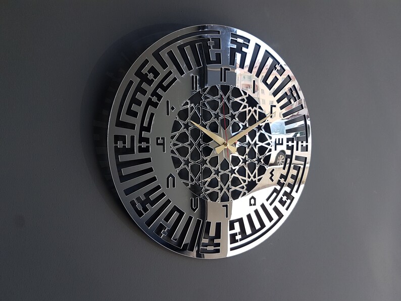 May include: A silver wall clock with black Arabic numerals and a geometric design in the center. The clock has a reflective surface and is hanging on a gray wall.