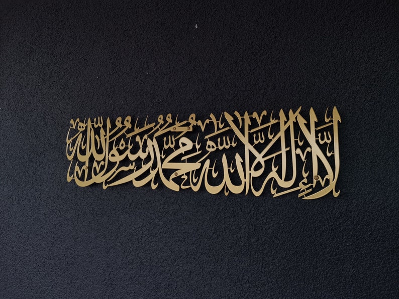 Może przedstawiać: Złotej barwy metalowa dekoracja ścienna z arabską kaligrafią na ciemnym, teksturowanym tle. Kaligrafia jest elementem dekoracyjnym, prawdopodobnie do dekoracji wnętrz lub cel&oacute;w duchowych.