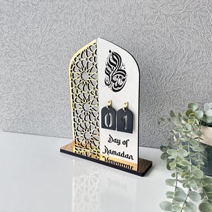 May include: A wooden Ramadan calendar with a gold and black geometric design. The calendar features a white background with black Arabic calligraphy and a black number "01" on a white tag. The text "Day of Ramadan" is printed below the number.