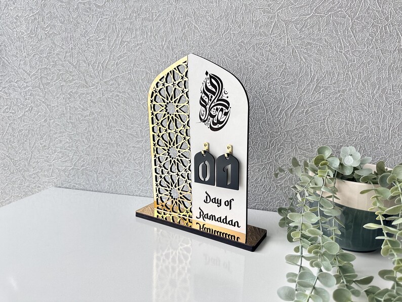May include: A wooden Ramadan calendar with a gold and black geometric design. The calendar features a white background with black Arabic calligraphy and a black number "01" on a white tag. The text "Day of Ramadan" is printed below the number.