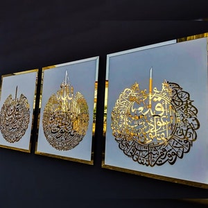 May include: Three framed wall art pieces with gold-colored Arabic calligraphy on a white background. The frames have a gold border. The artwork is displayed against a dark gray wall.