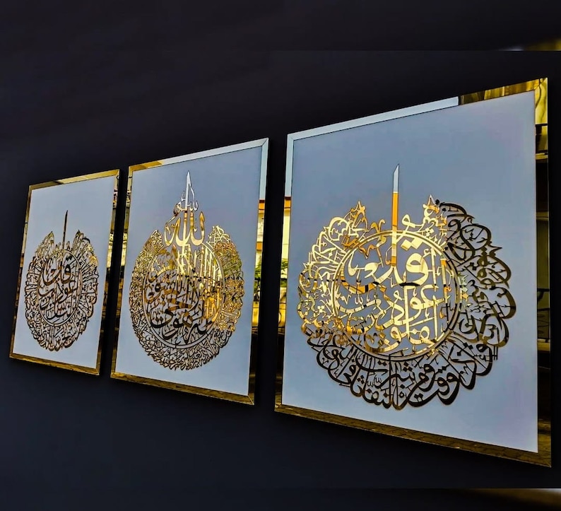 May include: Three framed wall art pieces with gold-colored Arabic calligraphy on a white background. The frames have a gold border. The artwork is displayed against a dark gray wall.