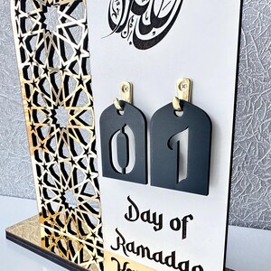 May include: A white wooden Ramadan countdown calendar with gold accents. The calendar features a black and gold geometric pattern, a black tag with the number "01" and the text "Day of Ramadan".
