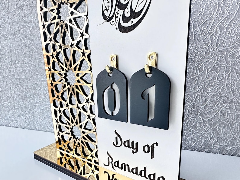 May include: A white wooden Ramadan countdown calendar with gold accents. The calendar features a black and gold geometric pattern, a black tag with the number "01" and the text "Day of Ramadan".