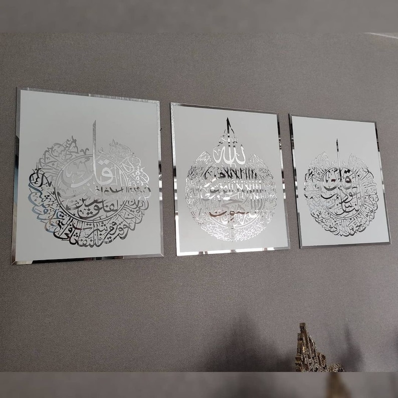 May include: Three framed wall art pieces with silver Arabic calligraphy on a white background. The calligraphy is intricate and detailed, with a reflective quality. The frames have a mirrored edge, adding to the elegant aesthetic.