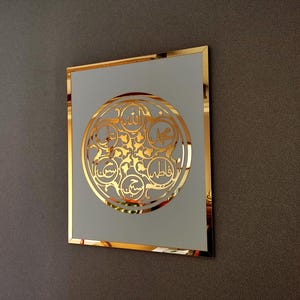 Könnte beinhalten: Ein quadratisches, goldgerahmtes Wandkunstwerk mit einem goldenen kreisförmigen Design. Das Design zeigt arabische Kalligraphie in einem dekorativen Muster. Der Hintergrund des Kunstwerks ist hellgrau. Die Wand ist dunkelgrau.