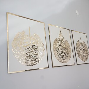 May include: Three framed Islamic calligraphy art pieces. Each features gold-colored script on a white background, framed with a gold border. The artwork is displayed on a light gray wall.