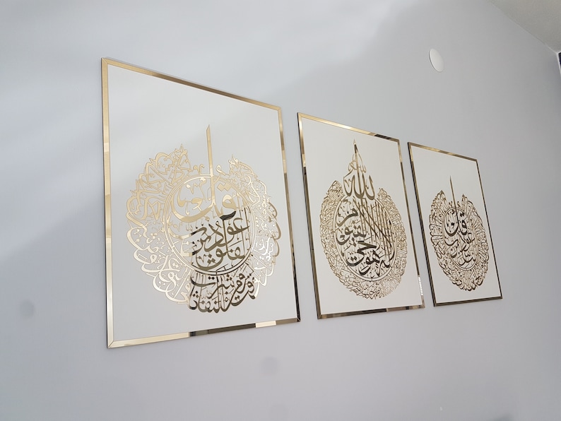 May include: Three framed Islamic calligraphy art pieces. Each features gold-colored script on a white background, framed with a gold border. The artwork is displayed on a light gray wall.