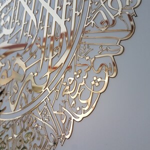May include: A gold-colored, ornate wall art piece featuring intricate Arabic calligraphy. The design is cut from a reflective material, creating a striking visual effect. The artwork is mounted on a light-colored background.
