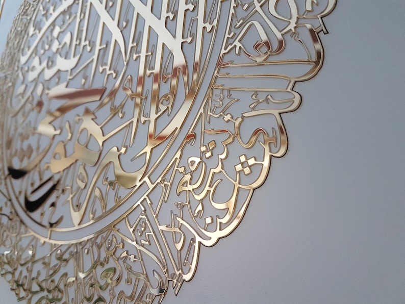 May include: A gold-colored, ornate wall art piece featuring intricate Arabic calligraphy. The design is cut from a reflective material, creating a striking visual effect. The artwork is mounted on a light-colored background.