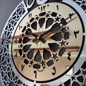May include: A silver and gold clock with a black face and Arabic numerals. The clock has a decorative design of stars and geometric shapes.