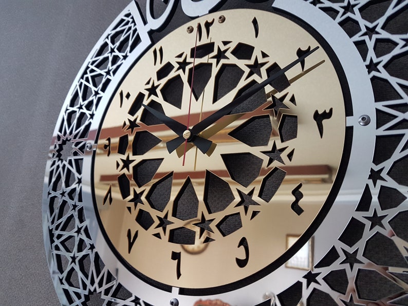 May include: A silver and gold clock with a black face and Arabic numerals. The clock has a decorative design of stars and geometric shapes.