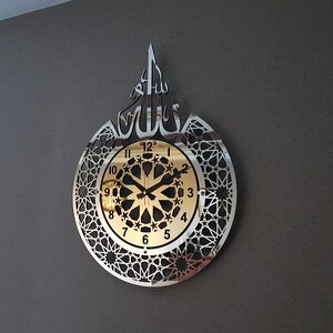 May include: A silver and gold wall clock with an intricate geometric design and the Arabic word "Allah" in the center. The clock face has a gold background with black numbers and hands.