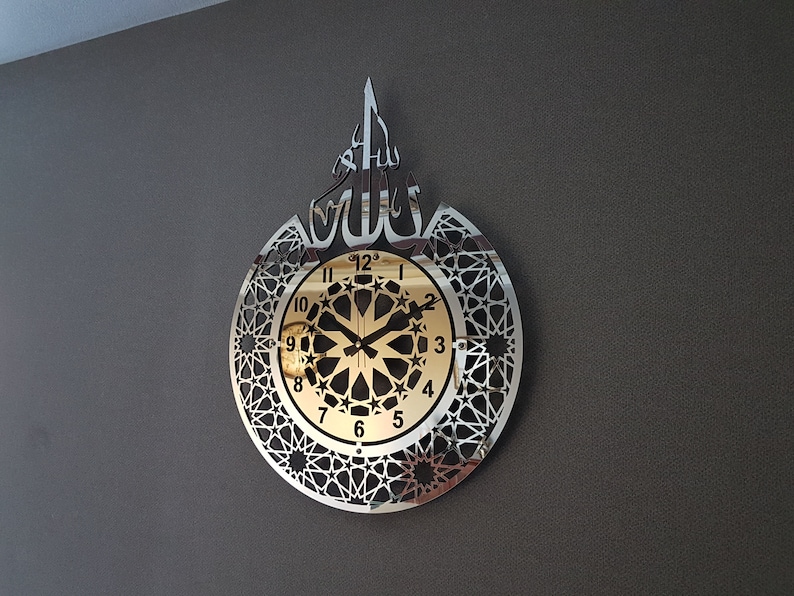 May include: A silver and gold wall clock with an intricate geometric design and the Arabic word "Allah" in the center. The clock face has a gold background with black numbers and hands.